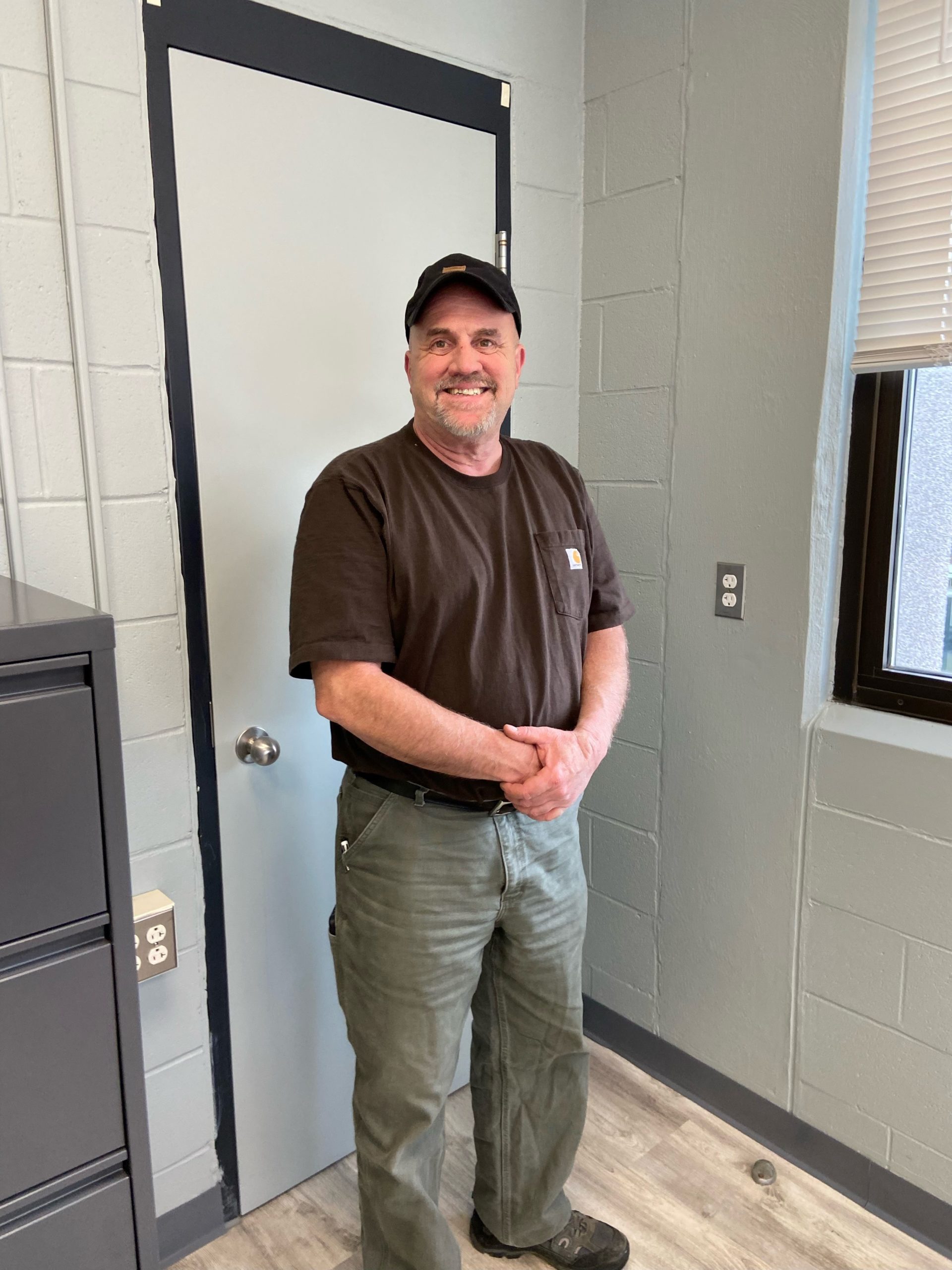 A man in a baseball hat and work clothes stands in front of a closed door.