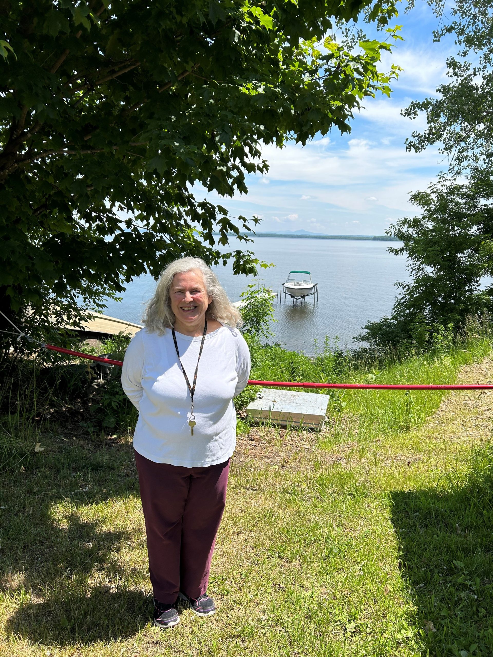 A woman with gray hair stands in front of a lake on a sunny day. There is a dock with a boat next to it in the distance.