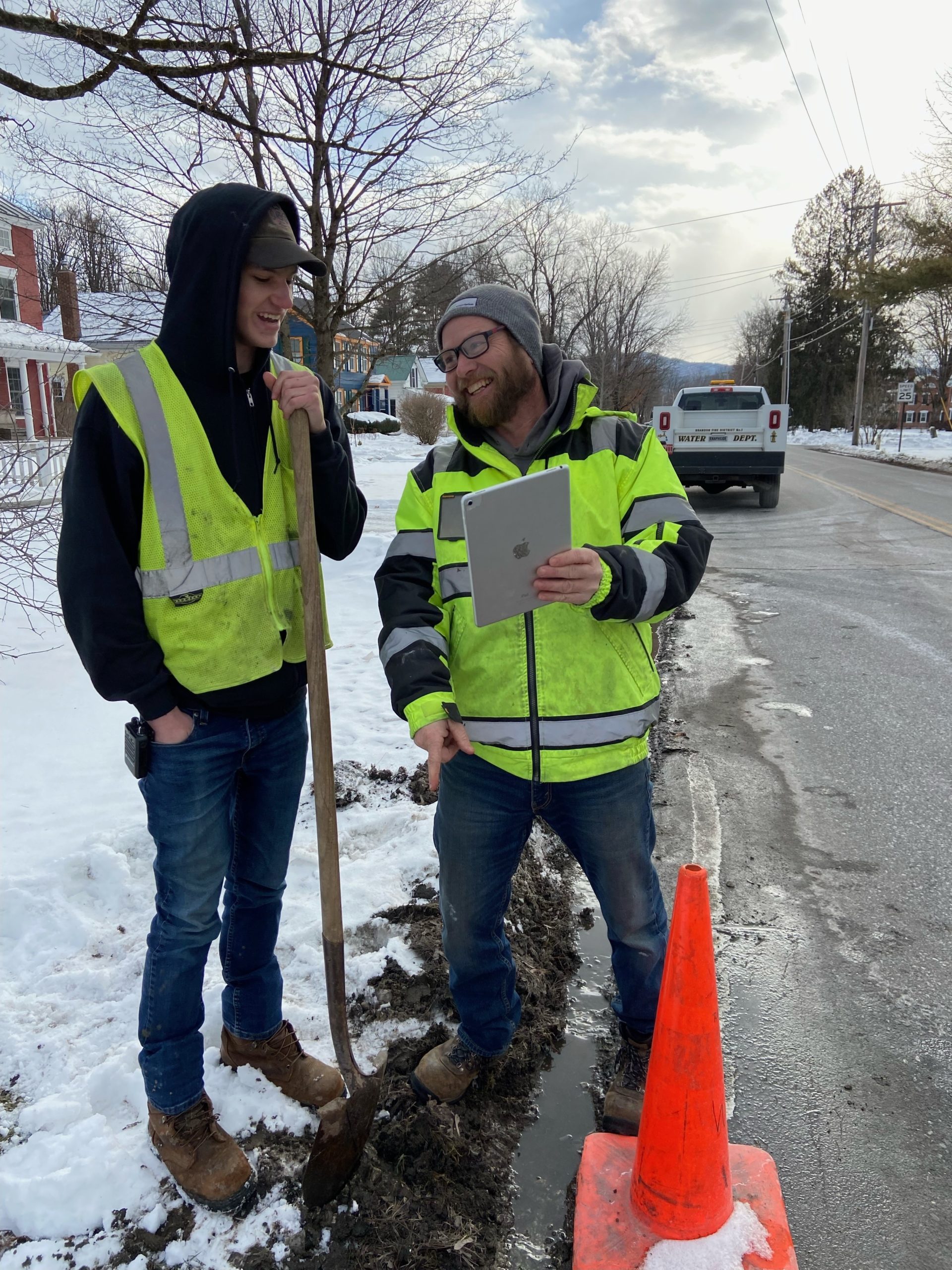 Two men stand on the side of a street. One holds a shovel and the other holds a tablet. There is snow on the ground.