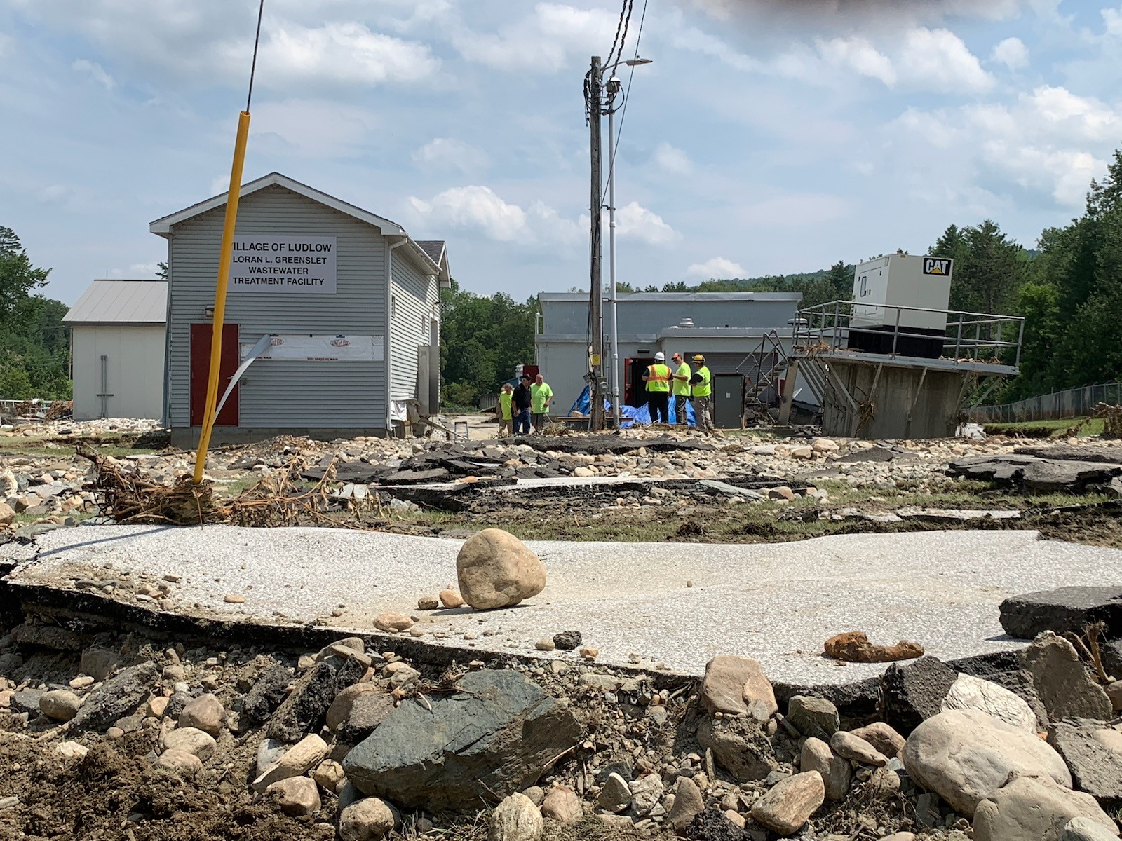 Rubble and rocks in the foreground. In the background, several people in safety vests stand next to a building with missing siding and a platform that is leaning slightly.