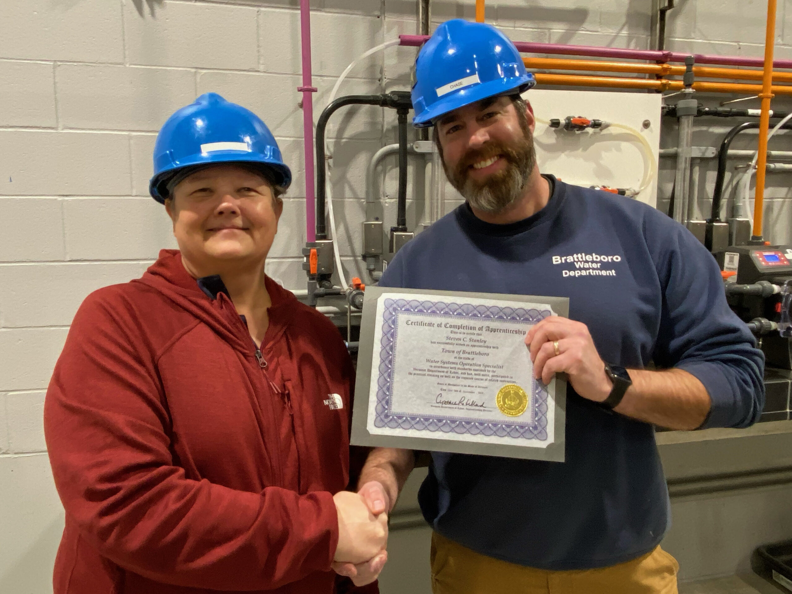 A man and a woman shake hands. Both are wearing blue hard hats. The man is holding a certificate.