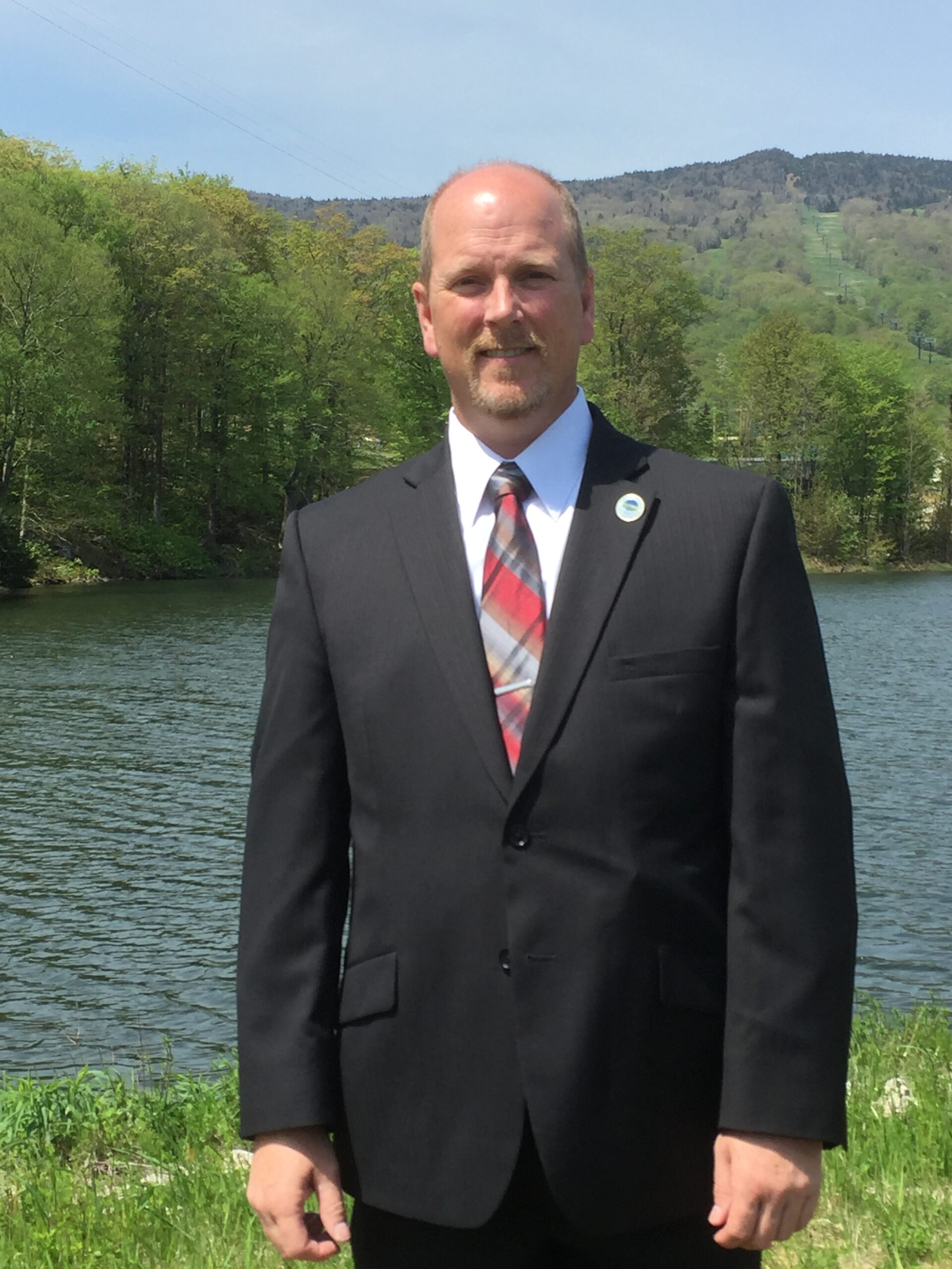 A man wearing a suit and tie stands in front of a water body with trees and a mountain in the background.