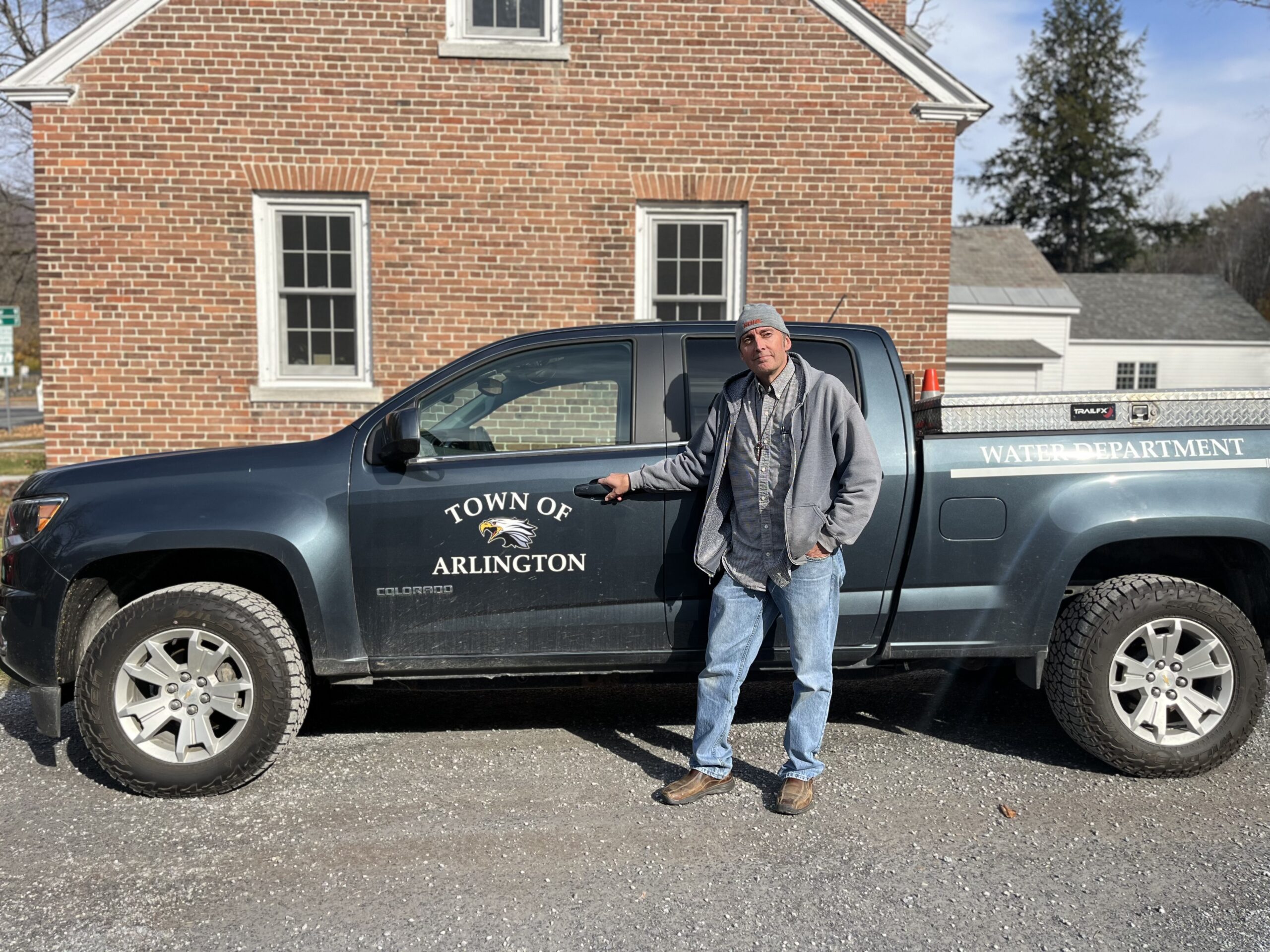 A man wearing a sweater and beanie stands next to a gray pickup truck that says "Town of Arlington"