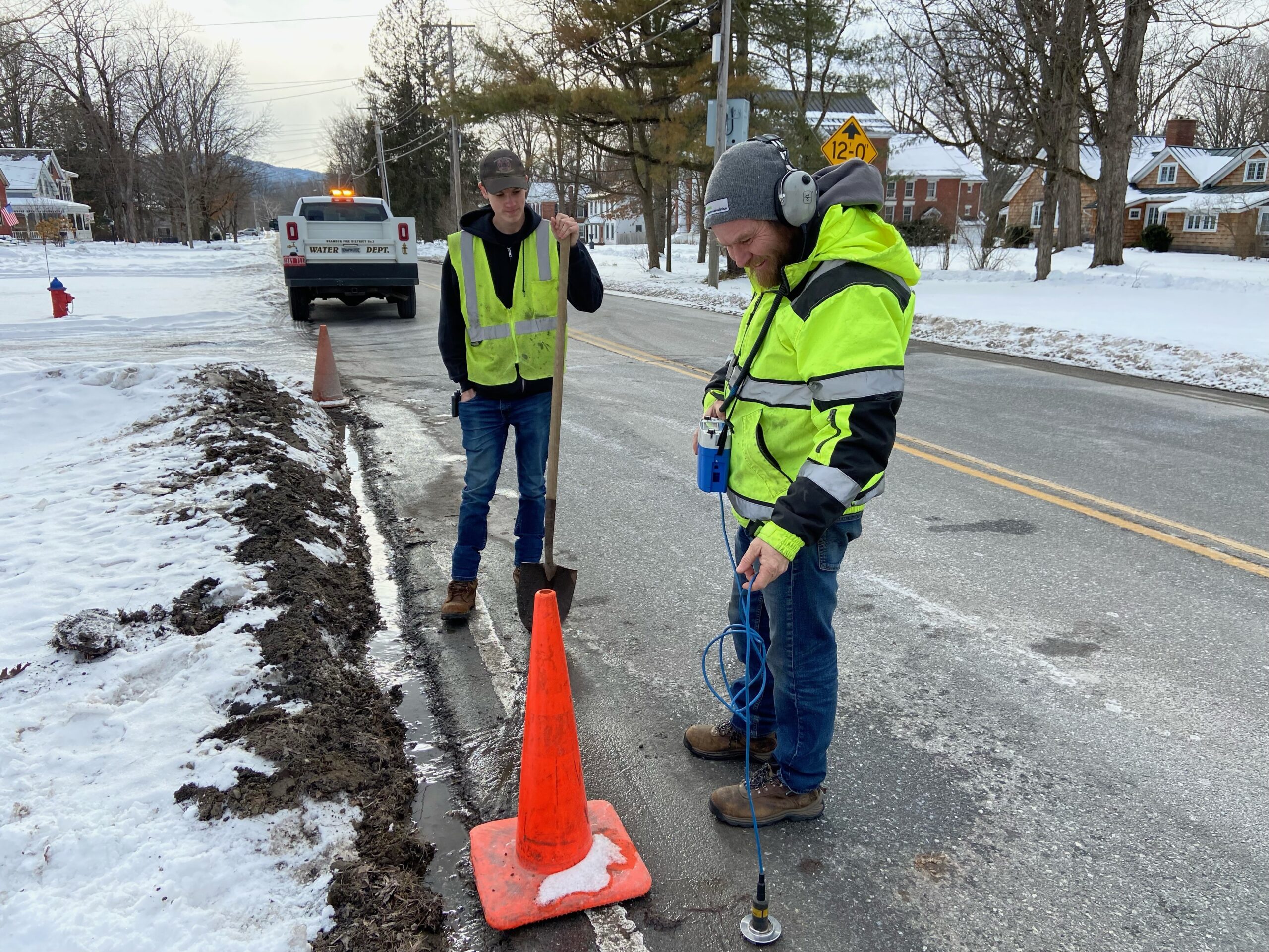 IMG_2178 To men in safety vests stand by the side of a road. There is water by the curb and a traffic cone. One man holds a shovel and the other is wearing headphones and holding a ground microphone to the asphalt.