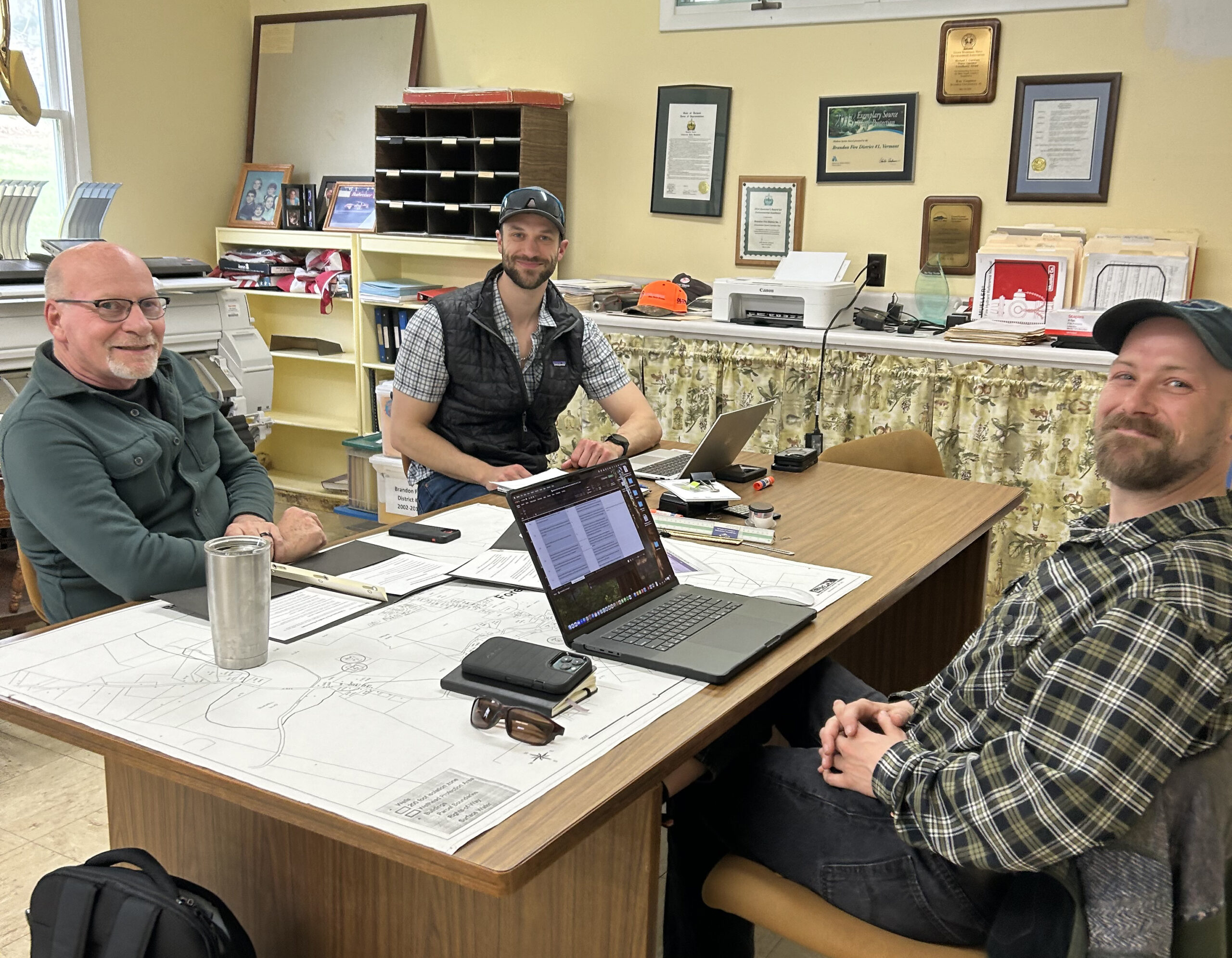 Three people sit around a desk with two laptops.