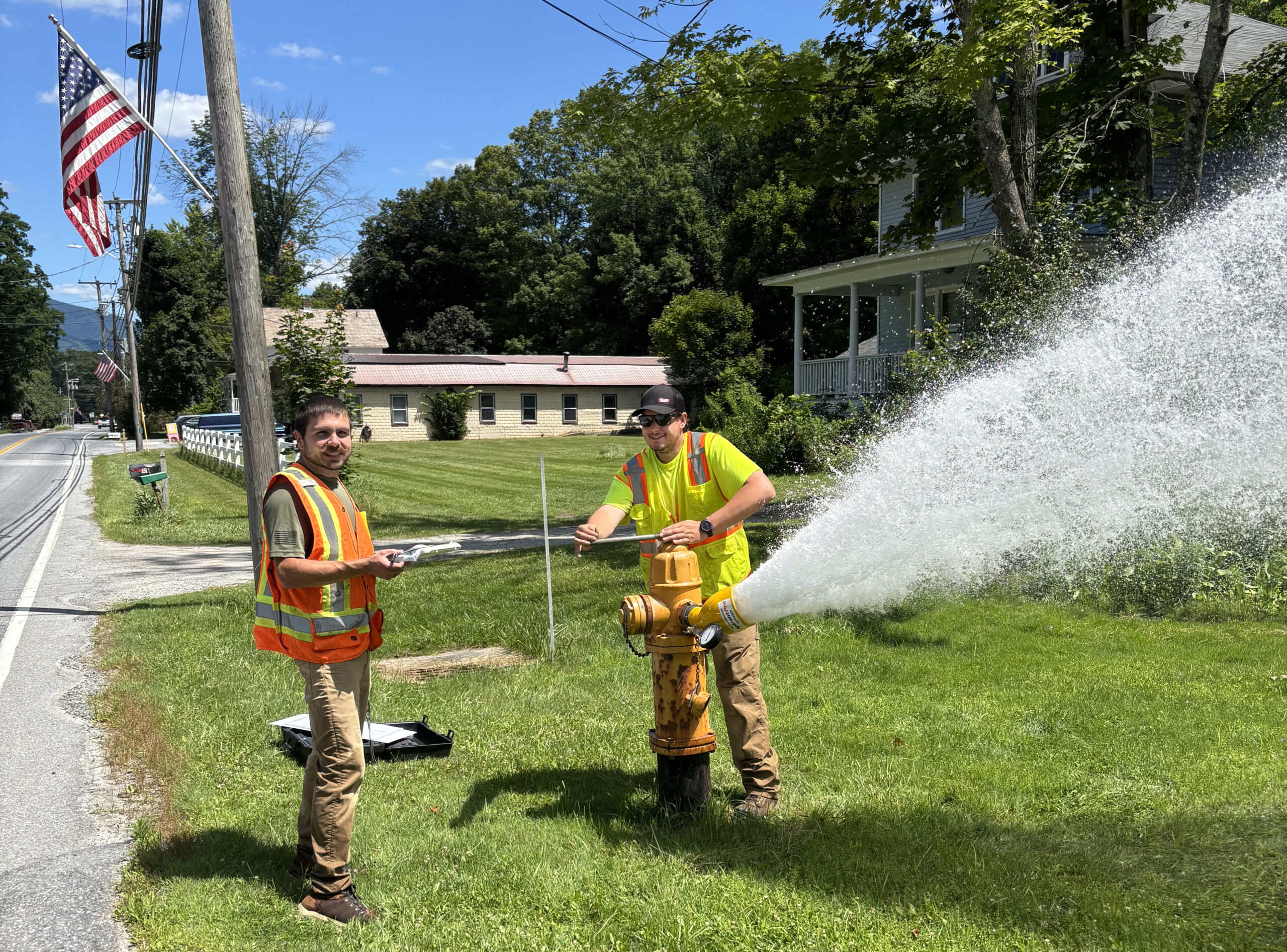 A man in a yellow safety vest holds a wrench on the top of a fire hydrant that is spraying water to the side. Another man in an orange safety vest holds a clipboard.