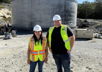 Two people wearing safety vests and hard hats stand in front of a concrete storage tank