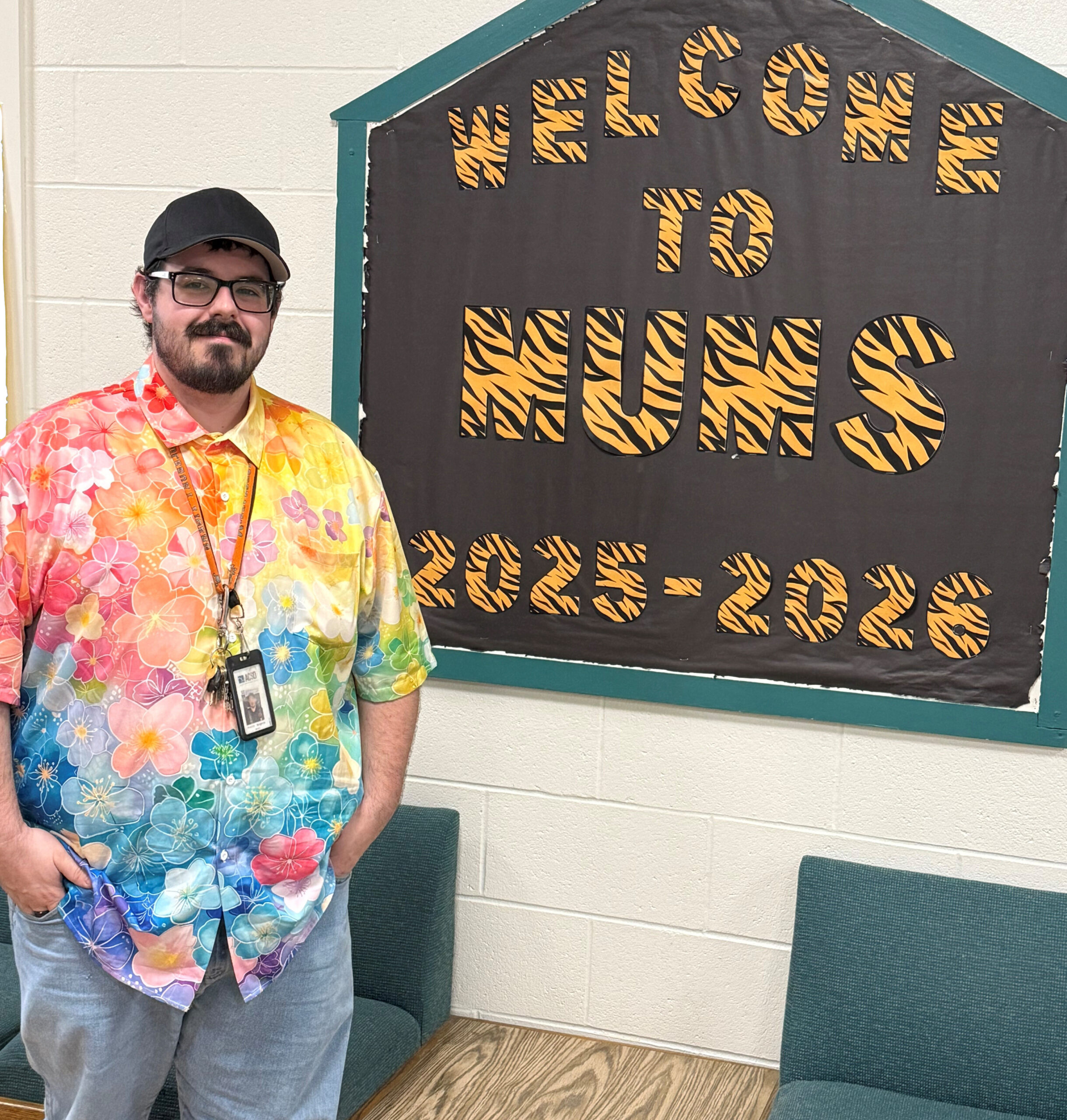 A man wearing a collared shirt with rainbow-colored flowers, a black baseball hat, glasses, and a lanyard stands in front of a bulletin board that reads "Welcome to MUMS 2025-2026"