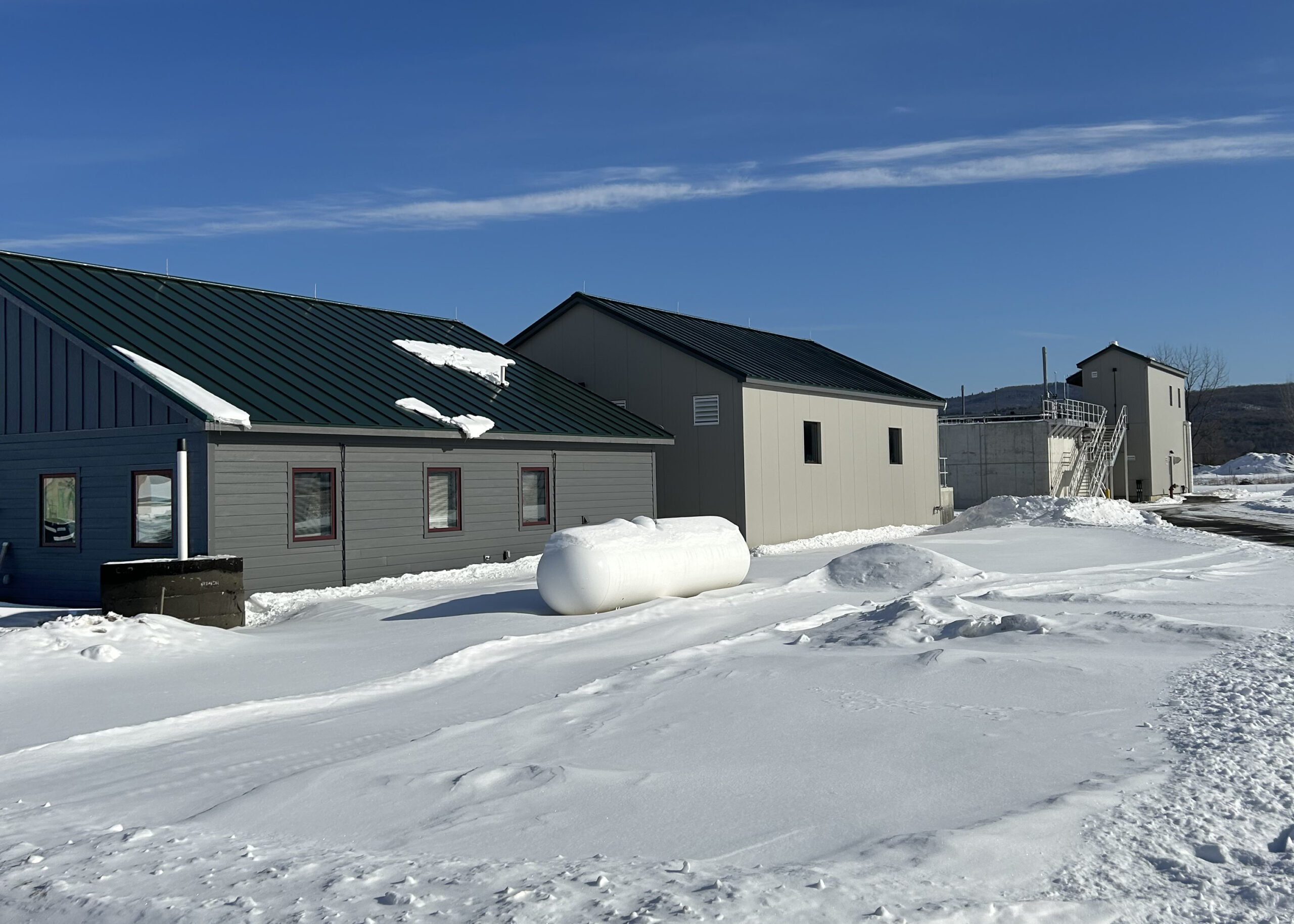 Several buildings stand in a row. There is a propane tank in front. The ground is covered in snow and the sky is blue.