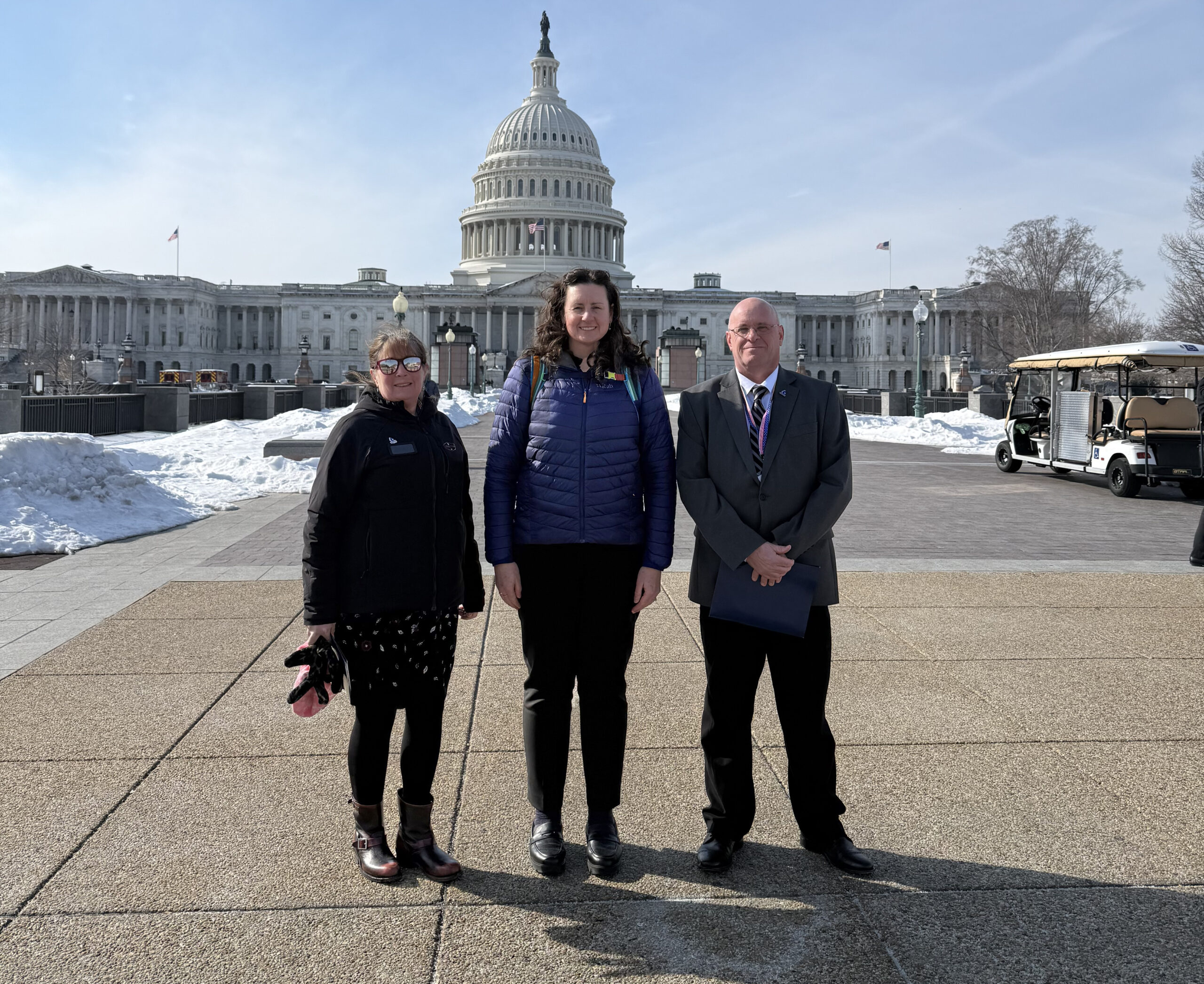 Three people stand together with the US Capitol Building in the background. There is snow on the ground and the sky is blue.