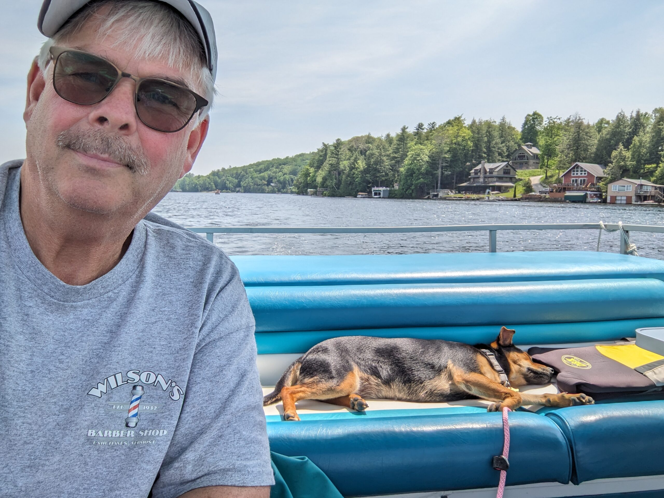 A man and a dog in a boat on a lake. There is a hill with trees in the background.
